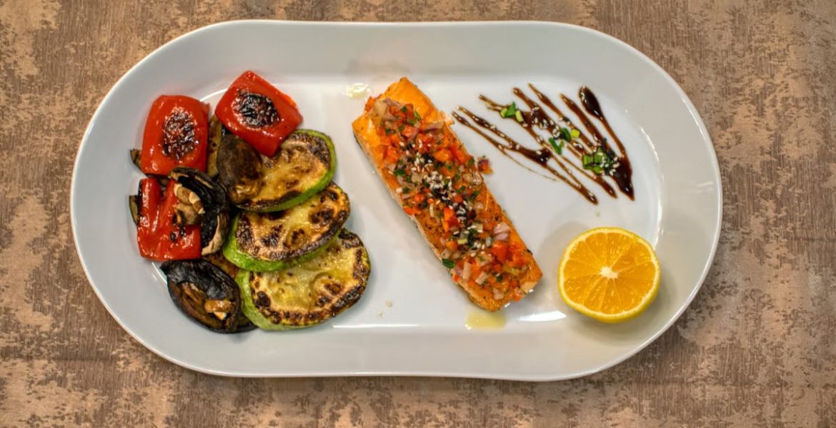 Gourmet grilled salmon served with roasted vegetables and lemon slice on a white plate.