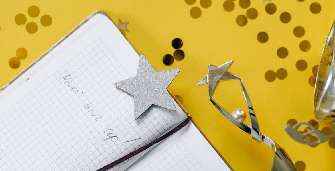 Flat lay of a notebook with motivational text, trophy, and star confetti on a yellow background.