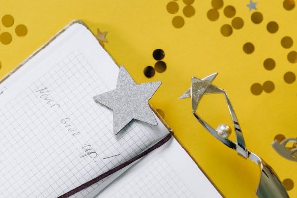 Flat lay of a notebook with motivational text, trophy, and star confetti on a yellow background.